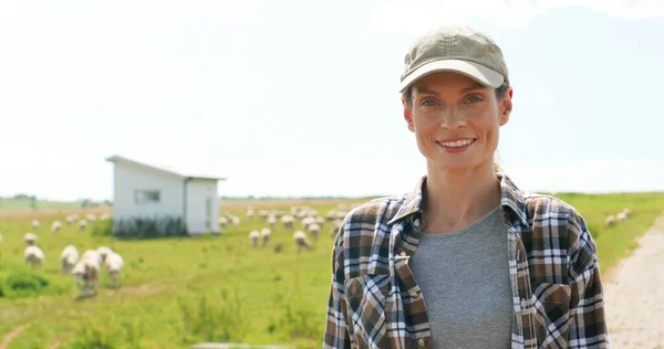 Portret van een mooie blanke jonge boerenvrouw met hoed op in het groene veld en glimlachend naar de camera. Schapen grazen op de achtergrond. Mooie vrouwelijke herder in de wei van vee. Platteland. — Stockfoto