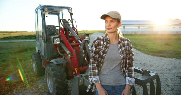 Açık havada büyük bir traktör makinesinin önünde duran ve kameraya neşeyle gülümseyen güzel beyaz kadın portresi. Çiftlikteki tarlada çalışan mutlu bir kadın çiftçi. Tarım. — Stok fotoğraf