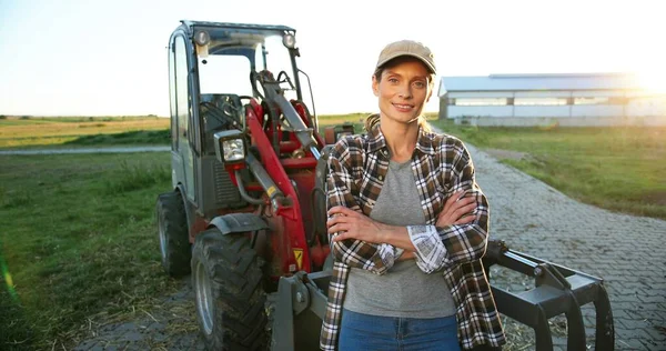 Portrait de belle jeune femme caucasienne en casquette debout à l'extérieur d'une grande machine tracteur et souriant joyeusement à la caméra. Jolie travailleuse agricole heureuse dans un champ à la ferme. Agriculture. — Photo