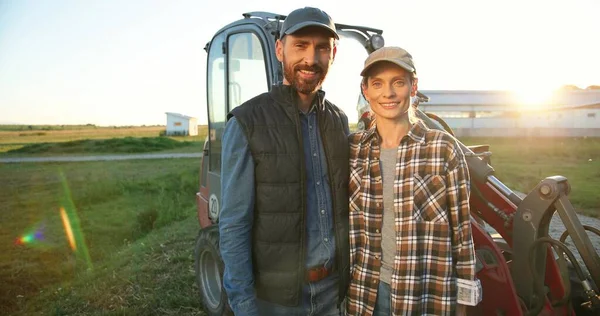 Portret Kaukasisch gelukkige echtpaar getrouwde boeren staan in knuffels op trekker, kijken elkaar aan en glimlachen naar de camera. Buiten. Een man die een vrouw omhelst. Landbouwconcept. Echtgenote en echtgenoot. — Stockfoto