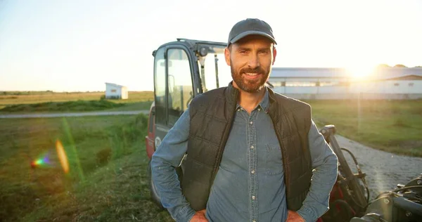 Portret van de jonge blanke knappe gelukkige boer die in het veld staat en glimlacht naar de camera. Grote tractor op de achtergrond. Vrolijke mannelijke werknemer op landbouwbedrijf. Zonlicht. Landbouw. — Stockfoto