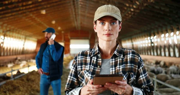 Jeune caucasienne heureuse jolie femme en chapeau à l'aide d'un appareil tablette et souriant dans l'écurie de la ferme. Portrait. agriculteur femelle tapant et défilant sur l'ordinateur gadget dans hangar. Homme parlant au téléphone en arrière-plan. — Photo