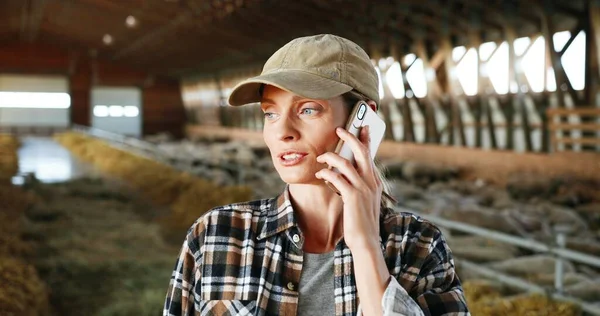 Gros plan de jeune femme blanche belle en casquette debout dans l'écurie avec troupeau de moutons et parlant sur téléphone mobile. Une agricultrice parle sur son portable dans un hangar à bétail. Conversation téléphonique. — Photo