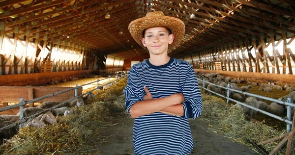 Portrait de petit garçon blanc en chapeau debout dans la remise avec troupeau de moutons et souriant à la caméra. Un petit adolescent dans une grange. L'été à la campagne. Enfance à la ferme animalière dans le village. Dolly shot. — Photo