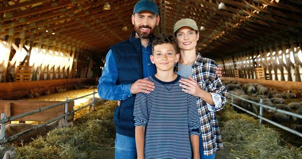 Portrait de mère et père heureux caucasiens avec un fils adolescent debout à l'écurie avec troupeau de moutons sur fond et souriant. Des parents joyeux avec un petit enfant dans une grange à la ferme. Dolly a tiré. Zoomer. — Photo