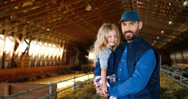 Portret van een blanke knappe vader die zijn handen vasthoudt aan een schattig dochtertje en glimlacht naar de camera bij schapenstal op de boerderij. Een boerenleven. Landelijk concept. Glimlachte man en klein meisje binnen. — Stockfoto