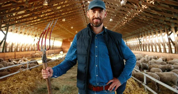 Portrait of young handsome Caucasian man farmer in cap looking at camera with pitchfork in hands and smiling in barn with livestock. Happy cheerful male shepherd in stable. Indoor Holding fork at farm — Stock Photo, Image