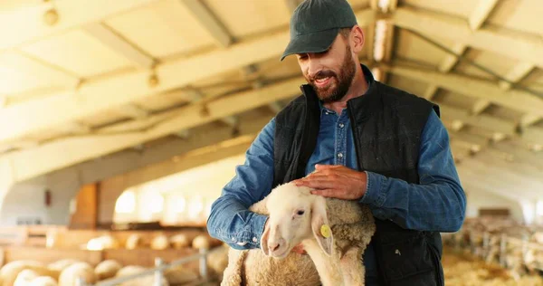 Portret van jonge knappe blanke man boer met pet lachend naar de camera en lammetje in de hand in schuur met vee. Gelukkig vrolijke mannelijke herder strelen schattig dier in stal. Peetschaap. — Stockfoto