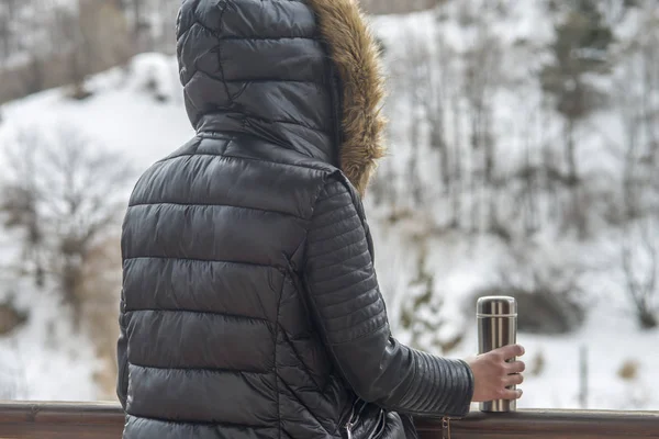 A girl in a winter jacket with a thermos of coffee or tea in her hand on the balcony in winter