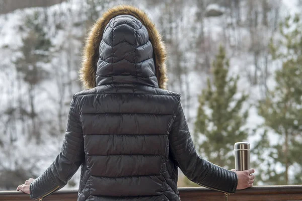 A girl in a winter jacket with a thermos of coffee or tea in her hand on the balcony in winter