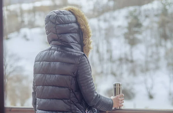 A girl in a winter jacket with a thermos of coffee or tea in her hand on the balcony in winter