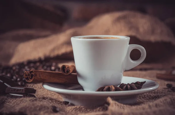 dark way of white cup of coffee with milk and aromatic spices: cinnamon sticks, star anise, dried slices of lemon and coffee beans on a napkin of burlap