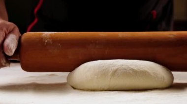 chef is rolling the dough with the rolling pin on the on the table. Hands close-up.Slow motion.Baker kneading dough for pizza  preparation. Chef cook making dough for baking pie on wooden table. Process preparation homemade pastry.food concept