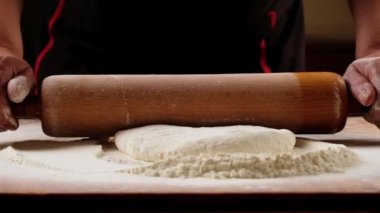 Sprinkled flour drops onto dough being rolled with a rolling pin by a chef. Slow motion.Baker kneading dough for pizza  preparation. Chef cook making dough for baking pie on wooden table. Process preparation homemade pastry.