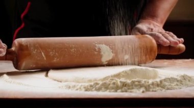 Sprinkled flour drops onto dough being rolled with a rolling pin by a chef.Slow motion.Baker kneading dough for pizza  preparation. Chef cook making dough for baking pie on wooden table. Process preparation homemade pastry.