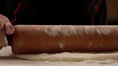 chef is rolling the dough with the rolling pin on the on the table. Hands close-up.Slow motion.Baker kneading dough for pizza  preparation. Chef cook making dough for baking pie on wooden table. Process preparation homemade pastry.