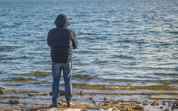 Одинокий человек стоит на пустом морском пляже и смотрит вдаль в холодный пасмурный осенний день, вид сзади.