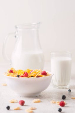 Bir kase mısır gevreği, sütlü, taze ahududu, yabanmersini. Sağlıklı kahvaltı için bir sürahi süt ve beyaz tahta üzerinde bir bardak.