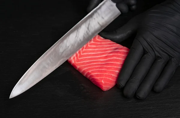 Chef cut fresh red salmon fish with sharp knife on black board . He is working on sashimi. Preparing traditional japanese sushi set. Only hands close up
