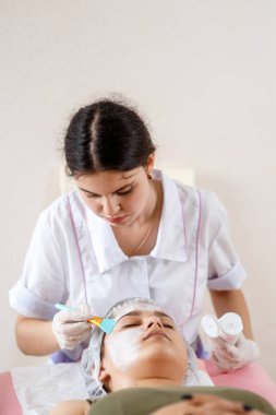 Kozmetik uzmanı yüz maskesi takıyor, fırça kullanıyor, deriyi sulu ve sağlıklı yapıyor. Çekici kadın dinleniyor. İşteki güzellik uzmanı. Kozmetiğin Elleri