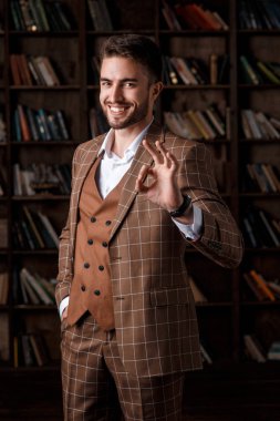 Kahverengi takım elbiseli sakallı genç adam kütüphanede dikilirken iyi bir hareket gösteriyor. İş adamı gülümsüyor ve sonuçtan memnun..