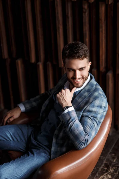 Jovem empresário de sucesso em um terno azul em uma gaiola está sentado em uma cadeira de couro. jovem com barba sorrindo. — Fotografia de Stock