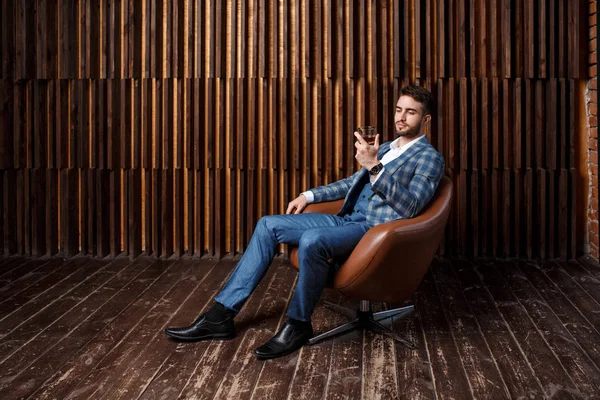 Odnoszący sukcesy brodaty biznesmen w niebieskim garniturze siedzi przy oknie w pokoju na poddaszu. Młody mężczyzna w garniturze pije whisky i koniak. — Zdjęcie stockowe