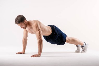 Spor fiziğinde sakallı genç bir adam beyaz tenli arka planda yerden şınav çekiyor, sporcu spor için içeri giriyor.