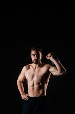 Siyah arka planda elinde kettlebell olan sakallı genç bir adam. Çıplak göğüslü sporcu
