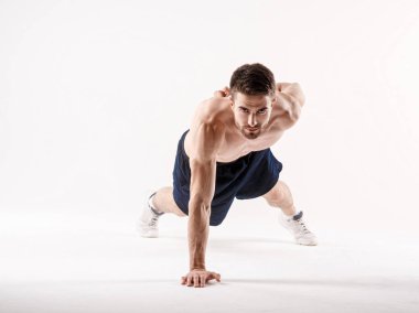 Spor fiziğinde sakallı genç bir adam beyaz tenli arka planda yerden şınav çekiyor, sporcu spor için içeri giriyor.