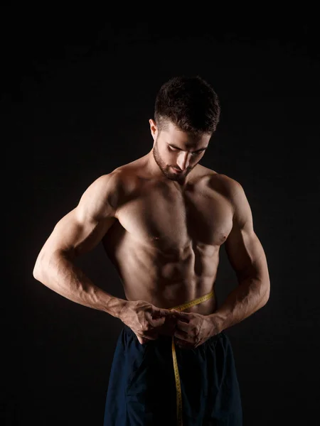 Bonito homem musculoso com fita métrica no fundo escuro. Conceito de perda de peso — Fotografia de Stock