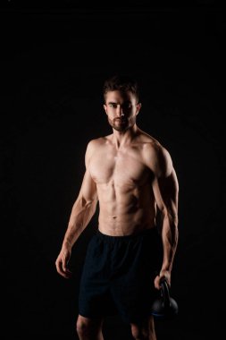 Siyah arka planda elinde kettlebell olan sakallı genç bir adam. Çıplak göğüslü sporcu