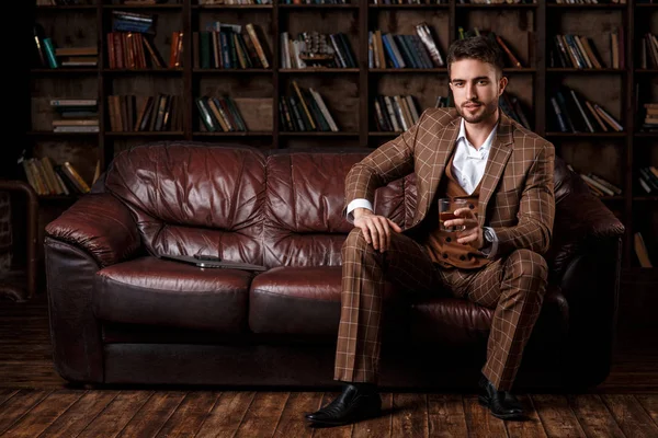Młody, odnoszący sukcesy mężczyzna w brązowym kosmtyum pije whisky. brodaty biznesmen siedzący w bibliotece na luksusowej skórzanej kanapie i pijący koniak — Zdjęcie stockowe