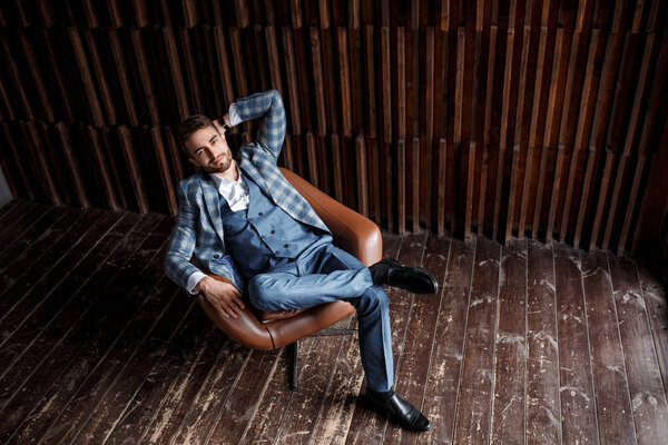 Молодой успешный бизнесмен в синем костюме в клетке сидит в кожаном кресле. молодой человек с бородой улыбается.