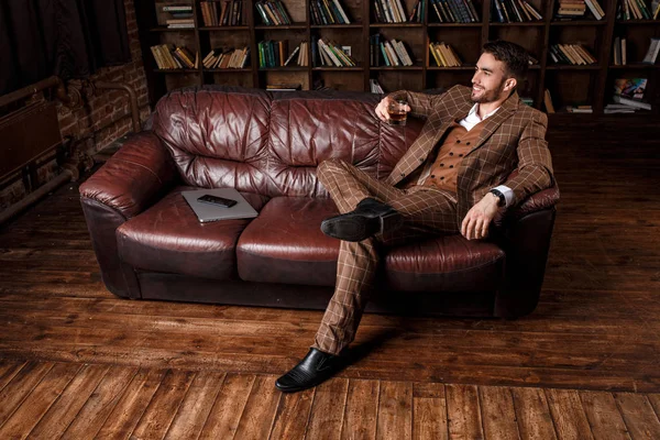 A fiatal, sikeres férfi barna kosmtyumban whiskyt iszik. szakállas üzletember ül egy könyvtárban egy luxus bőrkanapén és konyakot iszik. — Stock Fotó
