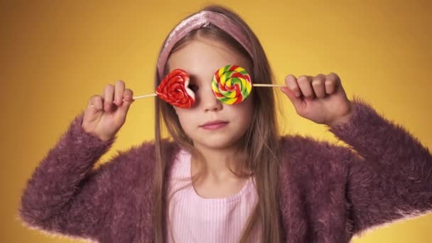 Sarı arka planda şeker taşıyan küçük bir kız. Çocuk elinde sopalarla sevinir ve güler.. — Stok video