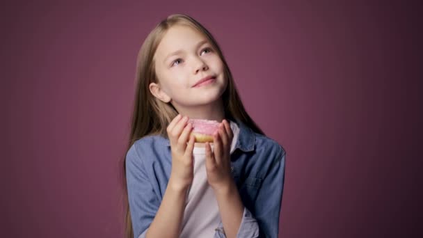 Gadis kecil makan donat dengan latar belakang merah muda. seorang anak menikmati permen — Stok Video