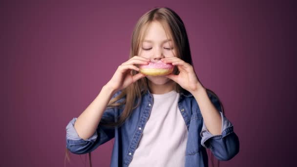 Ei lita jente spiser smultringer med rosa bakgrunn. et barn liker søtsaker – stockvideo