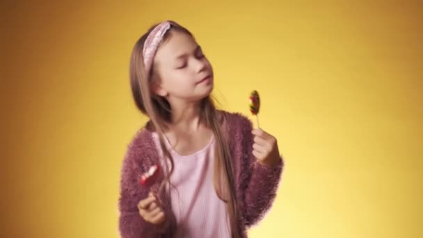 Menina com doces em um fundo amarelo. a criança se alegra e ri com paus em suas mãos. — Vídeo de Stock