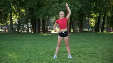 Sarı saçlı genç bir kız spor için doğaya gider. Sporcu parkta lastik bantlarla spor yapıyor.