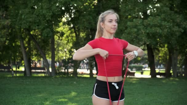 En ung flicka med blont hår går in för sport i naturen. idrottsman är engagerad i fitness med gummiband i parken — Stockvideo