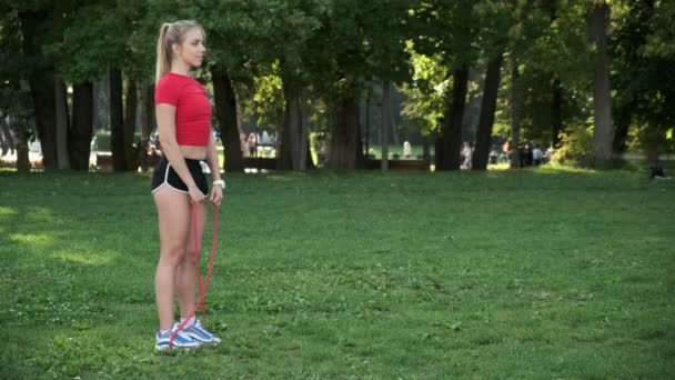Uma jovem com cabelo loiro entra para esportes na natureza. atleta está envolvido em fitness com elásticos no parque — Vídeo de Stock