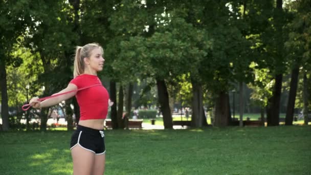Een jong meisje met blond haar gaat sporten in de natuur. atleet houdt zich bezig met fitness met elastiekjes in het park — Stockvideo