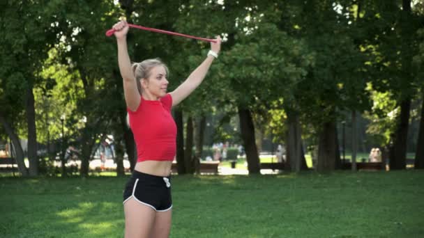 En ung flicka med blont hår går in för sport i naturen. idrottsman är engagerad i fitness med gummiband i parken — Stockvideo