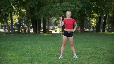 Sarı saçlı genç bir kız spor için doğaya gider. Sporcu parkta lastik bantlarla spor yapıyor.