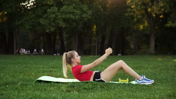 Eine junge Frau in rotem T-Shirt ist im Freien mit Fitness beschäftigt, das Mädchen macht in der Natur eine Übung auf dem Bauch. — Stockvideo