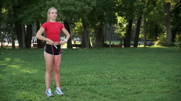 Ein junges Mädchen mit blonden Haaren treibt Sport in der Natur. Sportler treibt Fitness mit Gummibändern im Park — Stockvideo