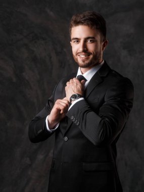 Gri bir arka plan üzerinde siyah takım elbiseli genç bir adam. Sakallı işadamı gülümsüyor