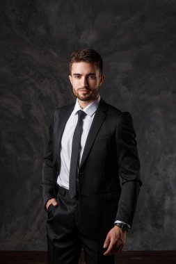 Gri bir arka plan üzerinde siyah takım elbiseli genç bir adam. Sakallı işadamı gülümsüyor