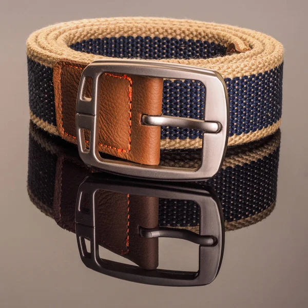 Rolled fashionable men\'s blue weaving belt with leather insert with silver matted metal buckle isolated on gray background. Studio shot on a dark background with reflection.